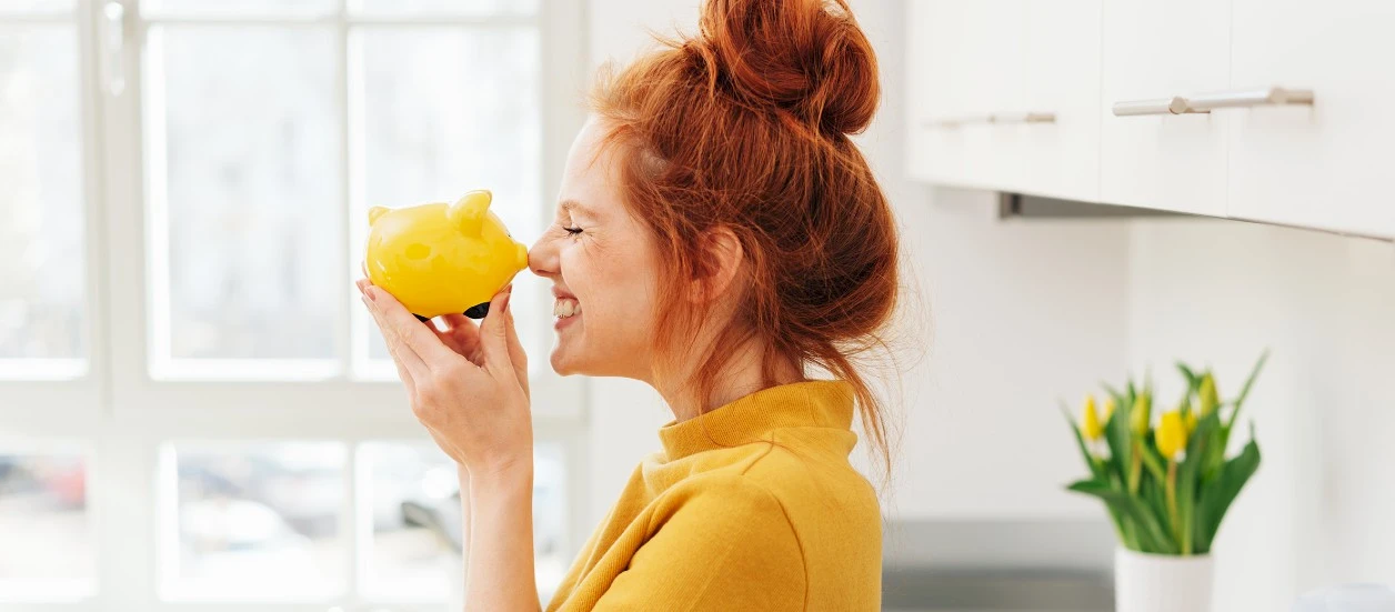 Lächelnde Frau die ein gelbes Sparschwein mit der Nasenspitze berührt. Unten links steht ‚Gewinnsparen‘ in grünem Kasten.“