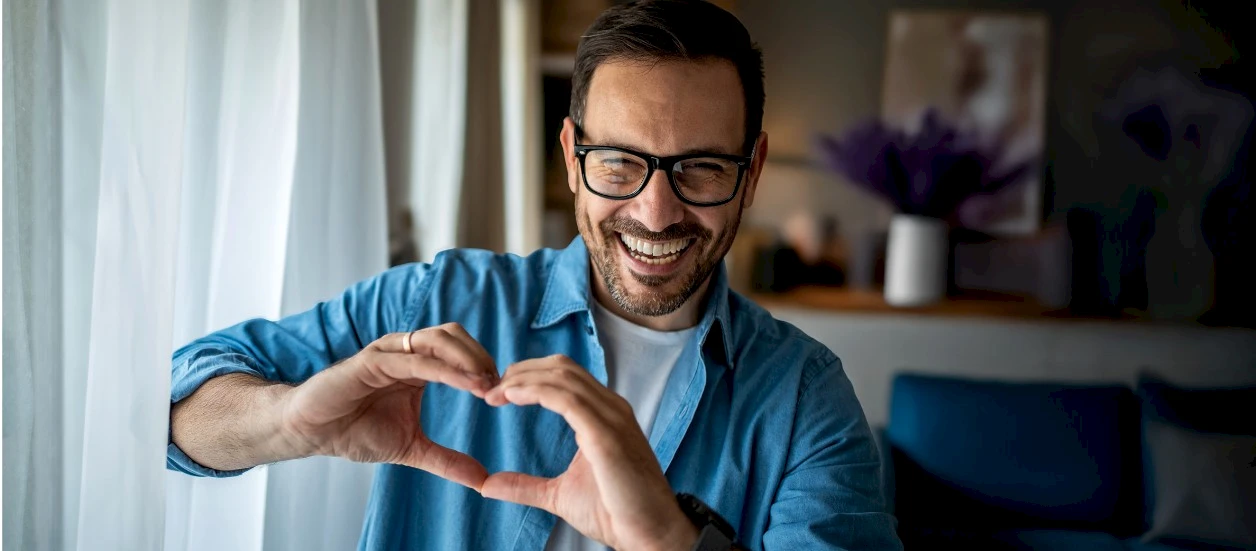 Mann mit Brille formt aus den Fingern ein Herz. Im Hintergrund steht eine Vase mit Lavendel.
