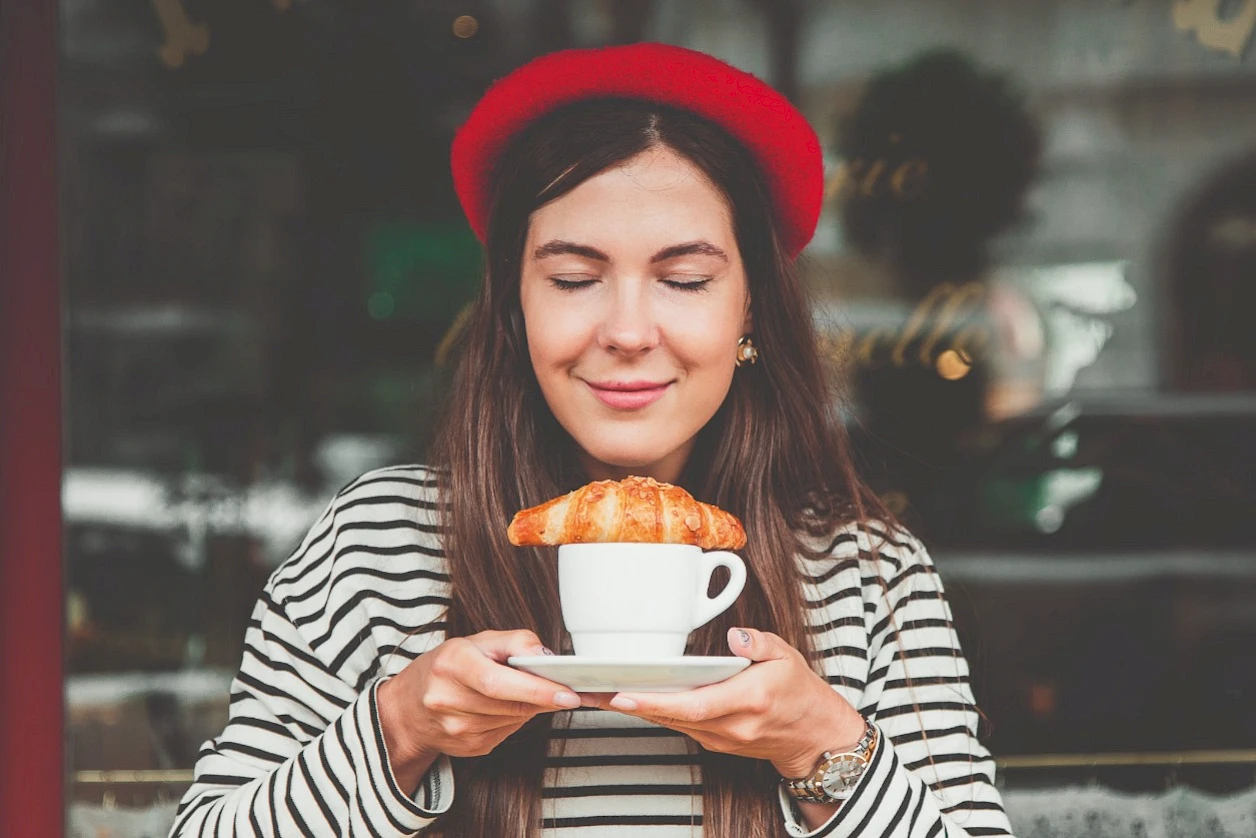 Junge Frau mit geschlossen Augen hält eine weiße Tasse mit einem Croissant darauf, trägt eine rote Baskenmütze und ein schwarz-weiß gestreiftes Oberteil, im Hintergrund ein Café.