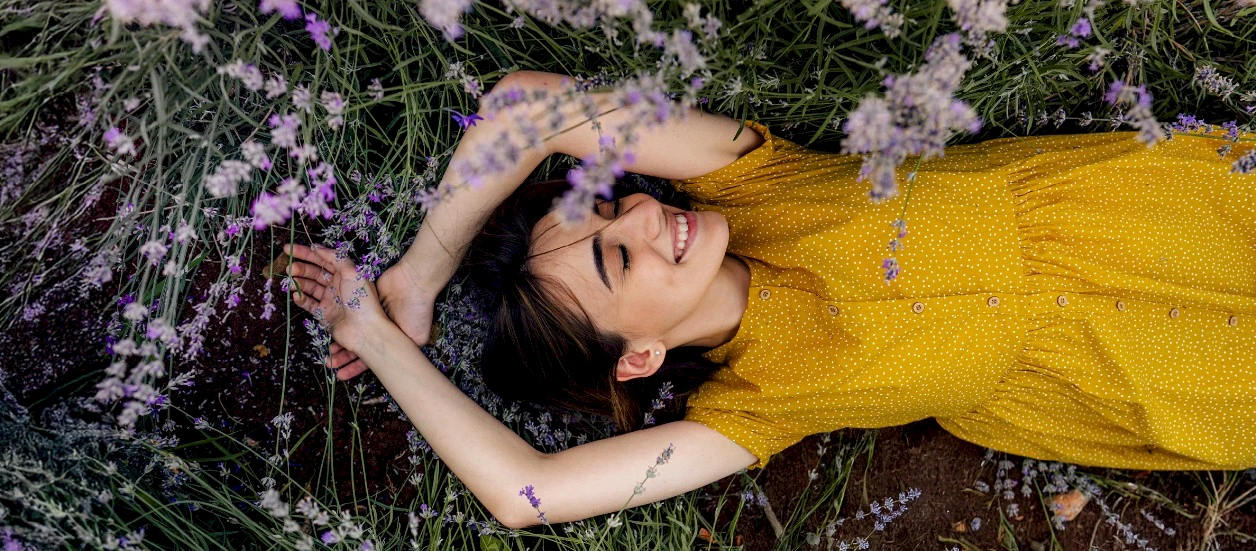 Frau in gelben Kleid liegt lächelnd mit geschlossenen Augen im Feld mit lila blühenden pflanzen.